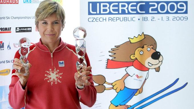 Prezidentka lyžařského šampionátu Kateřina Neumannová s oběma trofejemi před logem a obrázkem maskota lyžařského mistrovství.