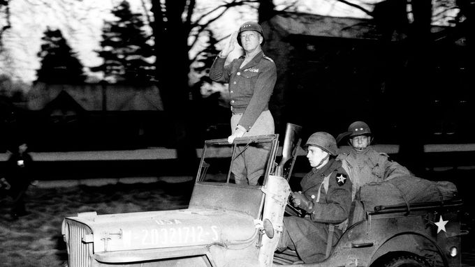Generál Patton (stojící) zdraví vojáky Druhé pěší divize při inspekci v severní Anglii v dubnu 1944. Na zadním sedadle velitel divize generálmajor Walter M. Robertson.