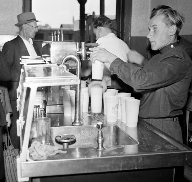 Červenec 1950. Dobový popisek: Družstvo cestující veřejnosti se stará, aby cestování na našich železnicích, lodích, na leteckých linkách bylo co nejpohodlnější a nejpříjemnější. V první vývojové etapě splnilo svůj plán zapojením všech nádražních restaurací, holíren a některých drobných pomocných zařízení na čs. železnici do družstva. Poté ministerstvo vnitřního obchodu pověřilo DCV, aby se ujalo dalších úkolů za združstevnění služeb na všech druzích čs. dopravy - u autobusové dopravy (ČSAD), letecké dopravy ČSA) i v paroplavební dopravě. U stánku s nápoji. (Pozn. red.: Bohužel nevíme, z jakého nádraží snímek pochází.) | Foto: ČTK