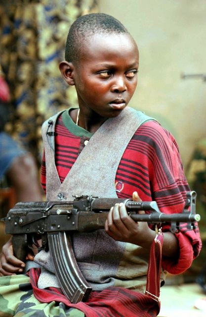 (dpa) - A boy soldier of the Union of Congolese Patriots (UPC) Roger, aged 12, watches passers by on the streets of Bunia, in the Democratic Republic of Congo, 10 June 2003. The Red Cross stated that fresh fighting in the town of Bunia, where a multinational peacekeeping force is being deployed, killed at least four people. Hundreds of militia fighters from the Lendu ethnic group attacked the ethnic Hema controlling the town of Bunia shortly after dawn on Saturday 7 June, a day after an advance party of European peacekeepers arrived. | Foto: ČTK