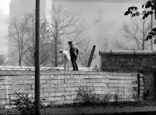 Berlínská zeď sice vyrostla v srpnu 1961, ale v průběhu následujících let byla neustále posilována a upevňována. Na snímku střeží východoněmecký příslušník bezpečnosti zedníka, který ji přizdívá. | Foto: ČTK