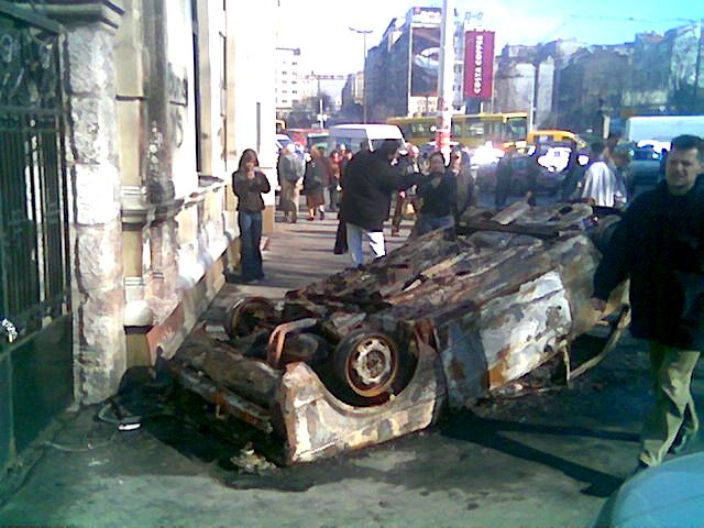 Převrácené ohořelé auto zůstalo po nočním řádění demonstrantů před vyrabovaným McDonaldem na Trgu Slavija v Bělehradě. (foto z mobilního telefonu) | Foto: Ondřej Besperát