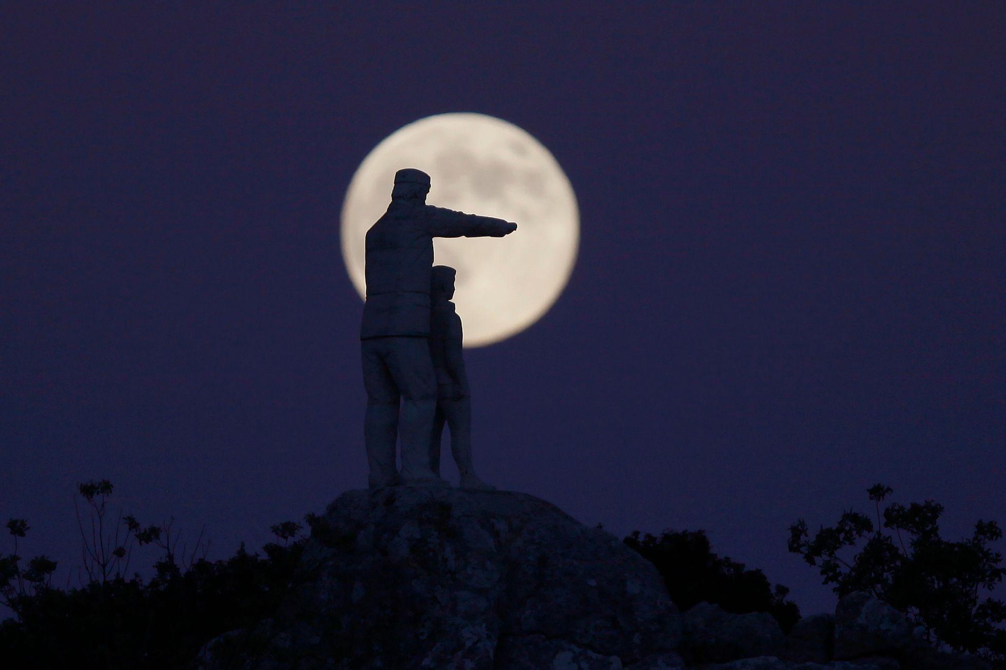 Statue of a man and a boy, is silhouetted against the supermoon as it rises at the Sierra de las Nieves nature park and biosphere reserve between El Burgo and Ronda