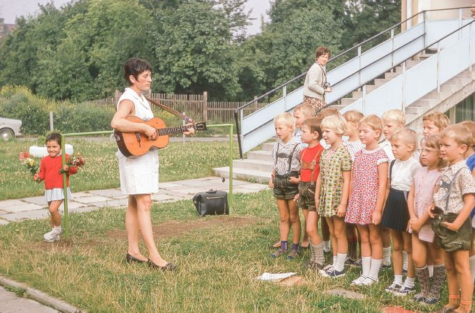 Skupina dětí poslouchá ženu hrající na kytaru před schodištěm, pravděpodobně v mateřské škole v NDR v 70. letech 20. století.