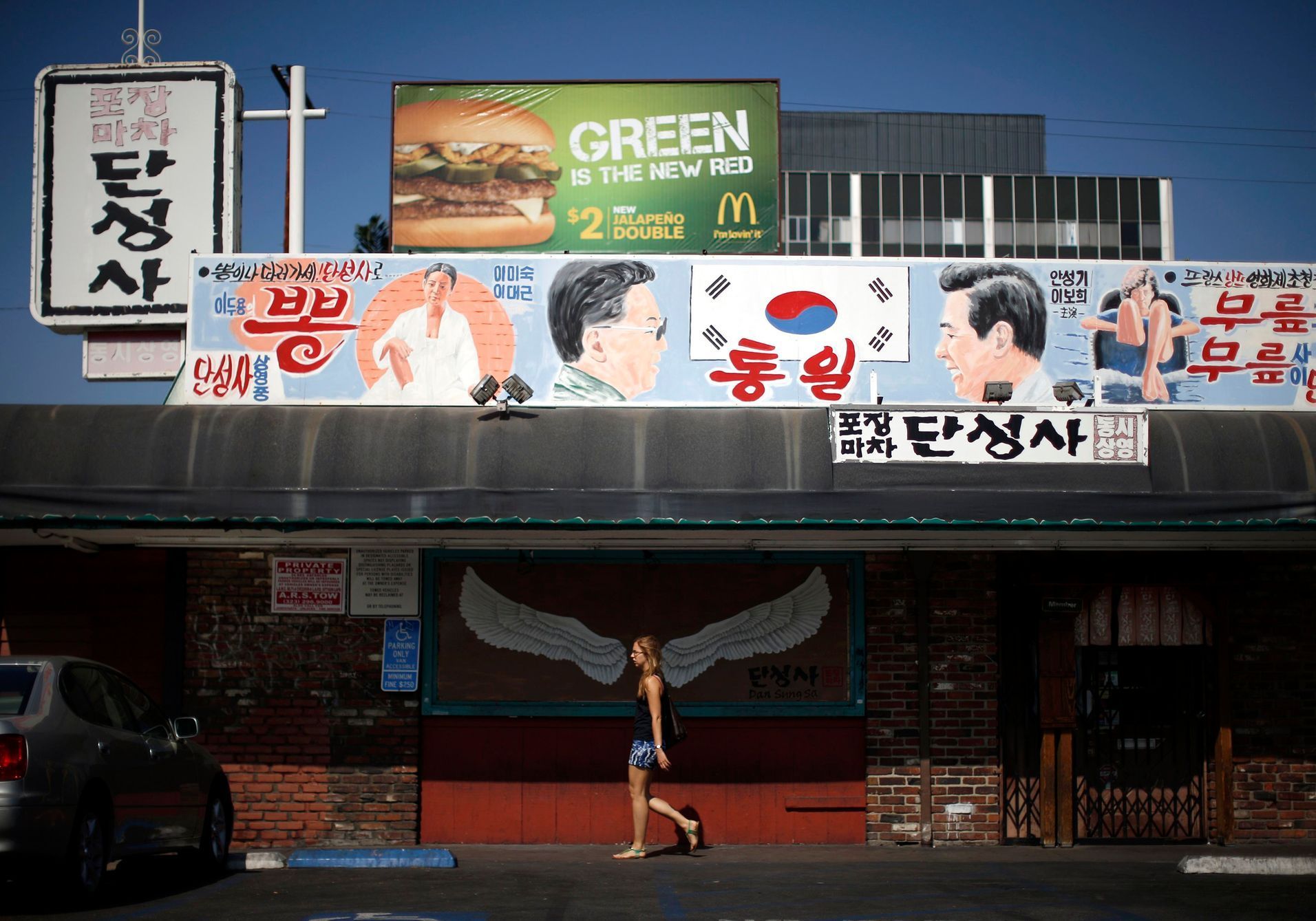 Woman walks past a mural of a pair of wings, under another mural depicting former North Korean leader Kim Jong Il and former South Korean president Kim Dae-jung, hanging above Dan Sung Sa restaurant a