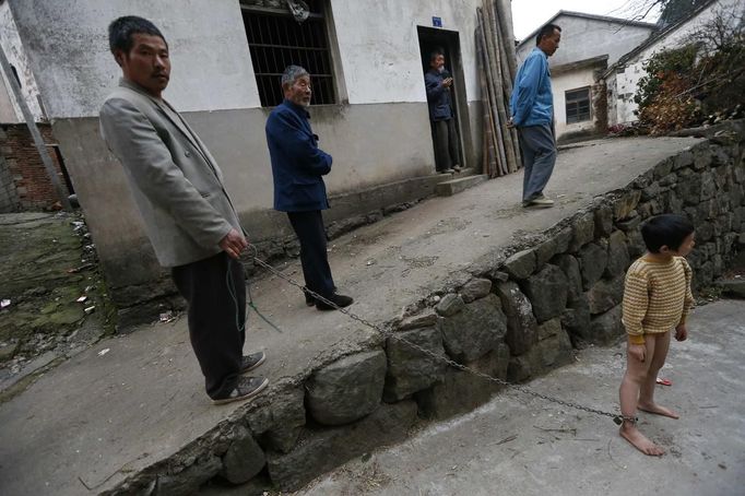 A boy stands outside his home as his father holds onto the chain locked around his ankle in Zhejiang province