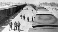 Květen 1944. Jeden z transportů je odbaven a čeká se na další "lidskou várku obětí". Stráže SS procházejí po příjezdové rampě tábora Auschwitz-Birkenau v Osvětimi. Rampa byla dokončena nedlouho před pořízením této fotografie. Deportační vlaky přijížděly dříve na rampu vzdálenou půl míle. Komíny krematorií II. a III. jsou vidět na horizontu.