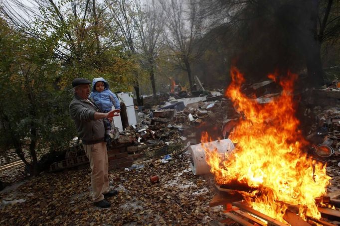 Justo Gabarri tries to keep his grandson Joel warm next to a bonfire as a bulldozer demolishes a home in the background in the Spanish gypsy settlement of Puerta de Hierro, in the outskirts of Madrid November 20, 2012. Fifty-four families have been living in Puerta de Hierro, on the banks of the Manzanares river for over 50 years. Since the summer of 2010, the community has been subject to evictions on the grounds that the dwellings are illegal. Families, whose homes have been demolished, move in with relatives whose houses still remain while the debris keeps piling up around them as more demolitions take place. Justo Gabarri's home was the second of two houses that got demolished Tuesday. REUTERS/Susana Vera (SPAIN - Tags: CIVIL UNREST BUSINESS CONSTRUCTION) Published: Lis. 20, 2012, 4:51 odp.