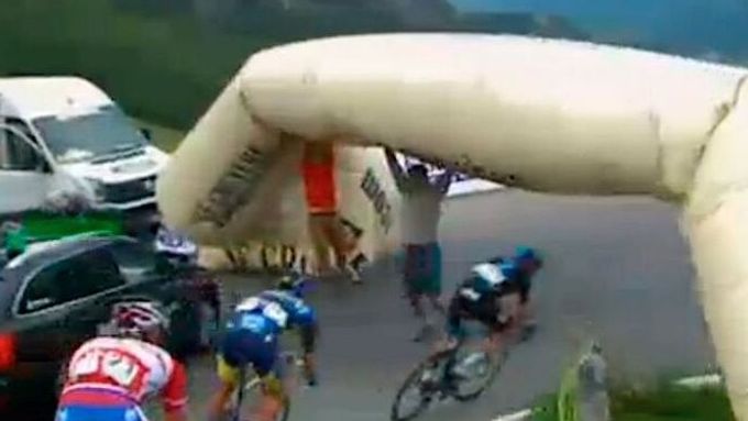 Cyklisté podjíždějí spadlý reklamní banner, který drží pořadatelé a diváci.