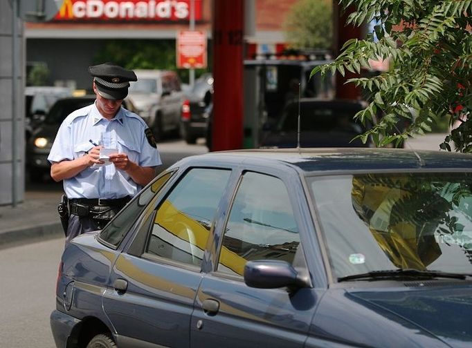 Policista uděluje pokutu řidiči zeleného Escortu za parkování v těsné blízkosti křižovatky a přechodu pro chodce.