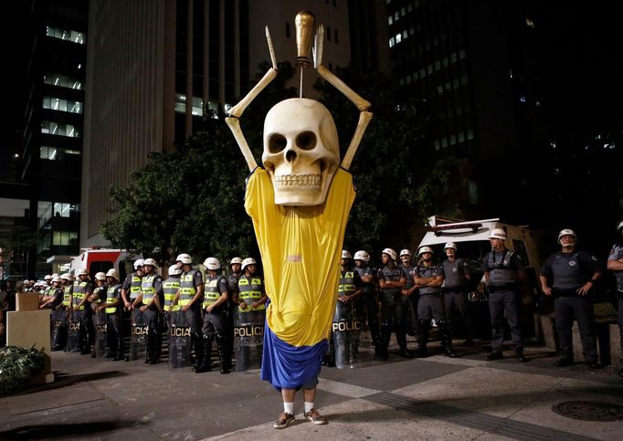 Military policemen stand in a line behind a demonstrator wearing the figure of a skeleton holding up a trophy representing that of the FIFA World Cup during a protest aga