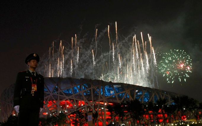 Policista stojí na stráži. Za ním září ohňostroj olympijského ceremoniálu nad Národním stadionem v Pekingu.