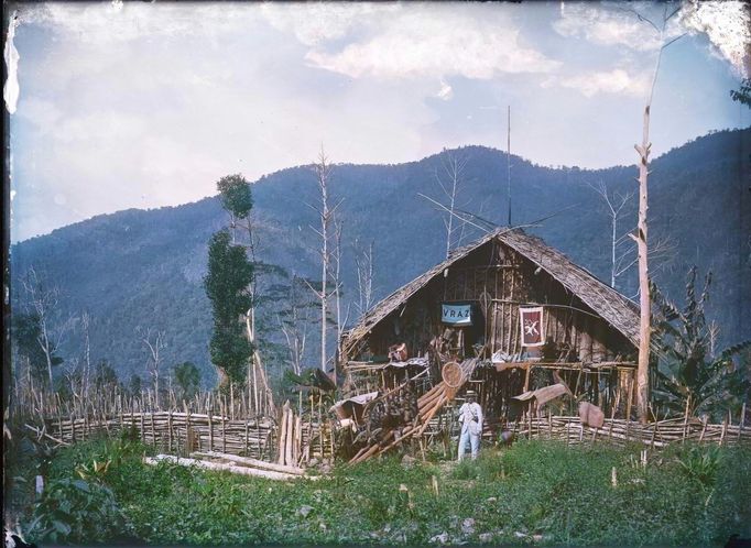 Enrique Stanko Vráz se síťkou na chytání hmyzu před obytnou chýší označenou vlajkou s českým lvem a nápisem Vraz. Pohoří Hattam. Nová Guinea, 1896.  Kolorovaný snímek