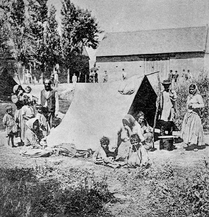 Moravští cikáni, fotografie Romů u stanu, na pozadí stromy a stodola, historická pohlednice, kolem roku 1890.