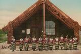 Maorští tanečníci před vyřezávaným domem ve vesnici Ohinemutu u jezera Rotorua. V době, kdy vznikla tato fotografie, tedy mezi lety 1890 až 1910, bylo Ohinemutu hlavním centrem celého okolního regionu.