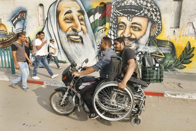 Wheelchair-bound Palestinian freelance photographer Moamen Qreiqea holds his wheelchair as he rides on a motorcycle past murals of late leader Yasser Arafat (R) and late Hamas spiritual leader Ahmed Yassin, on a street in Gaza City October 1, 2012. Qreiqea, 25, lost both his legs in an Israeli air strike in 2008 while taking pictures east of Gaza. The father of two is determined to continue his career despite his disability. REUTERS/Suhaib Salem (GAZA - Tags: SOCIETY MEDIA) Published: Říj. 1, 2012, 3:39 odp.