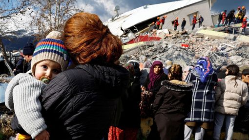 Žena drží dítě a stojí u trosek a škod po zemětřesení v tureckém Gaziantepu.