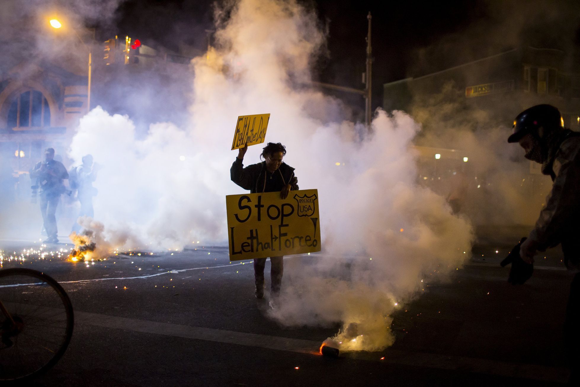 A protester holds a sign as clouds of smoke and crowd control agents rise, shortly after the deadline for a city-wide curfew passed in Baltimore, Maryland