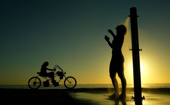 Žena si dává po plavání sprchu, v pozadí projíždí muž na kole a slunce zapadá nad pláží Manly Beach v Sydney, 25. prosince 2003. O Vánocích bylo tehdy 27 stupňů, surfaři, plavci a cyklisté začínali okupovat pláže již kolem půl šesté hodiny ráno. REUTERS/Tim Wimborne