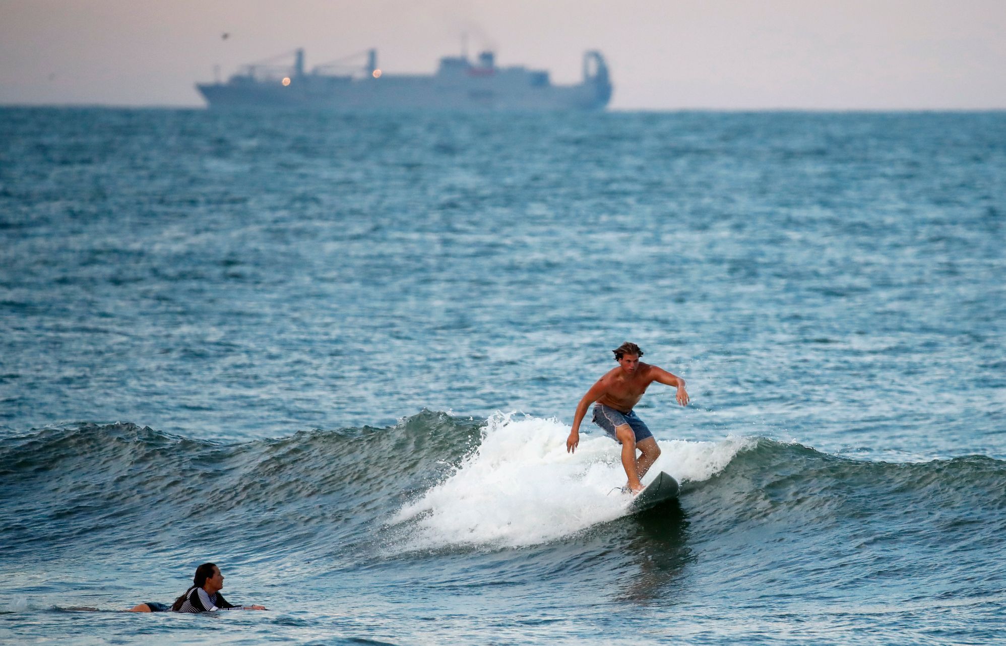 A surfer catches a wave with a navy ship behind him, Tuesday, Sept. 11, 2018, in Virginia Beach, Va., before the arrival of Hurricane Florence.