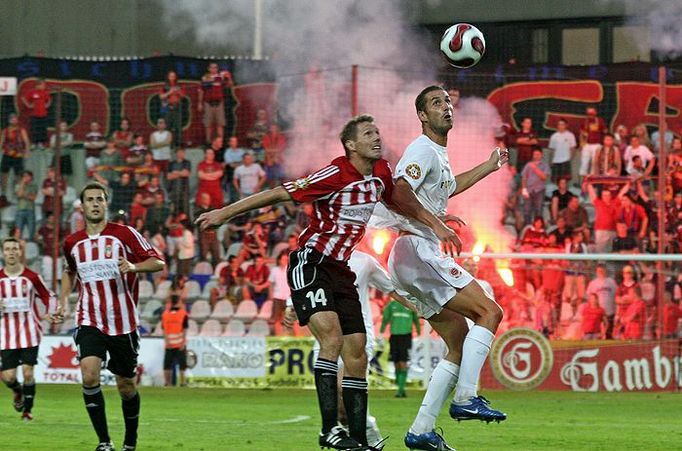 Libor Došek (AC Sparta) a Tomáš Kučera (FK Viktoria Žižkov) v boji o míč, zatímco fanoušci Sparty už slaví vítězství zapálenými světlicemi.