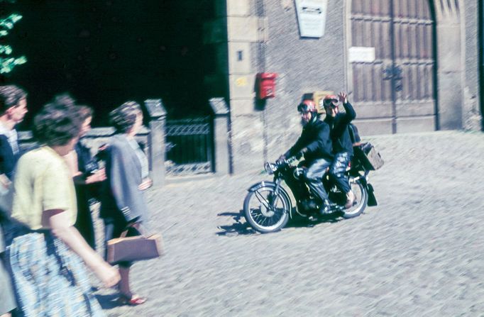 Dva muži na motocyklu projíždějí kolem skupiny přihlížejících v městském prostředí v NDR v roce 1959. Lokace neuvedena.