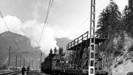 "Na elektrifikaci tratě Žilina - Spišská Nová Ves se začalo pracovat slavnostním výkopem ve Varíne roku 1949. Stavba od té doby úspěšně pokračovala, takže v první polovině června uvedou do provozu úsek Štrba-Poprad. Na ostatních úsecích jsou práce v takovém stavu, že koncem tohoto roku bude elektrifikovaná trať i v úseku Žilina - Spišská Nová Ves. Na jednotlivých úsecích probíhají dokončovací práce. Pracovní čety vyvíjejí obětavou činnost a pomocí socialistického soutěžení zrychlují elektrifikaci. K jakým výsledkům vede socialistické soutěžení je vidět i na časech montáže trakčního vedení jednoho kotevního pole, tj. cca 1200 metrů. Nejlepší je četa vedená náčelníkem Zoltánek Mihálym na úseku Vrútky-Rožumberok, která provedla montáž jednoho kotevního pole mezistaničního úseku za 6,03 výlukových hodin, což je téměř rekord. Na druhém místě je oddíl náčelníka Ondreje Kerdíka, jehož členové smontovali jedno kotevní pole mezistaničního úseku za 6,28 výlukových hodin. Na třetím místě je četa náčelníka Jozefa Pelikána, která ukončila montáž za 6,31 výlukových hodin, přičemž průměrný čas je 7 hodin. Na úseku Liptovský Mikuláš-Štrba v těchto dnech probíhají zkušební jízdy elektrických lokomotiv s přepravou osobních a nákladních vlaků. Ukazuje se, že dvě elektrické lokomotivy nahradí výkonem 3 parní při stejné zátěži. Elektrifikovaná doprava je hospodárnější, rychlejší i bezpečnější. V osobní dopravě znamená příjemnější, pohodlnější a hygieničtější cestování. Na snímku: pracovní četa, vedená náčelníkem oddílu Zoltánem Mihálym, pracovala při rozvíjení trolejového drátu na úseku Vrútky-Ružomberok s pomocí zvláštního vlaku." Dobový text k fotografii pořízené koncem května 1955.