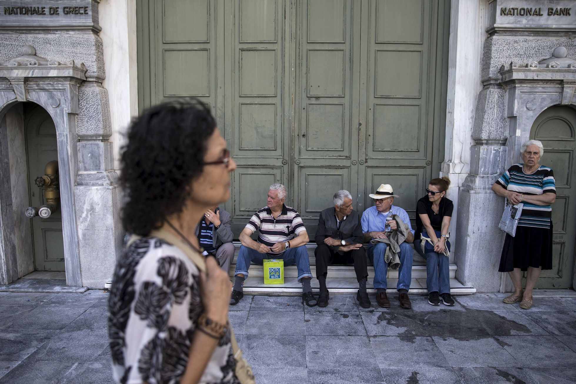 People, most of them pensioners, sit outside a closed National Bank branch at the bank's headquarters in Athens