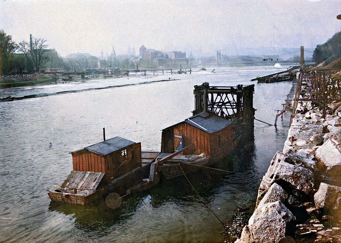 Pohled na drtič skály u Holešovického břehu během splavnění Vltavy u ostrova Štvanice. V pozadí most císaře Františka Josefa I.. Rok cca 1909. Kolorováno.