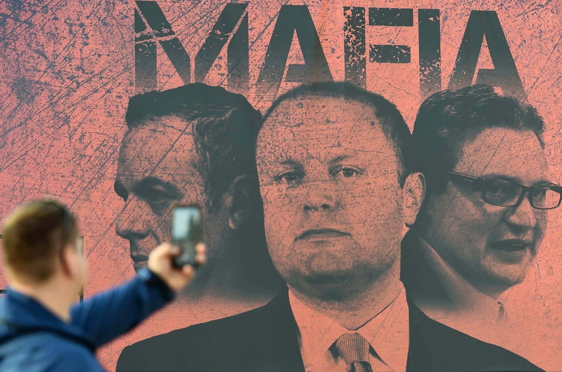 Muž fotografuje plakát, zobrazující maltského premiéra Josepha Muscata a jeho dva ministry jako mafii.