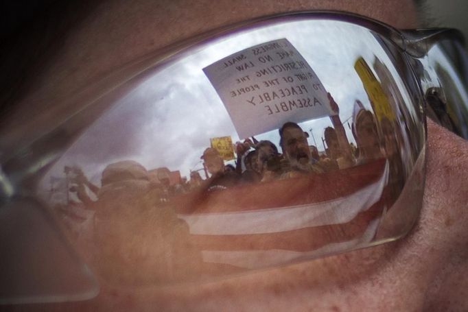 Demonstrators holding the U.S. Flag and a placard are reflected in the glasses of a police officer blocking protesters marching near the site of the Democratic National Convention in Charlotte, North Carolina on September 4, 2012. President Barack Obama will be nominated as the Democratic candidate at the convention, along with Vice President Joe Biden as his running mate, for November's U.S. presidential election. The placard in the reflection reads, "Congress shall make no law restricting the right of the people to peacefully assemble." REUTERS/Adrees Latif (UNITED STATES - Tags: POLITICS ELECTIONS CIVIL UNREST TPX IMAGES OF THE DAY) Published: Zář. 4, 2012, 10:29 odp.