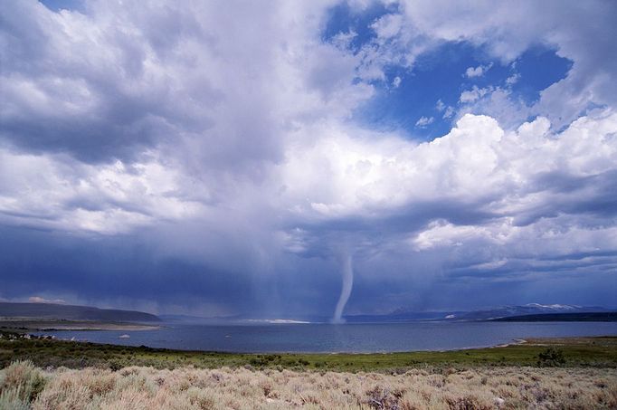 A storm and tornado form over water in a desert landscape