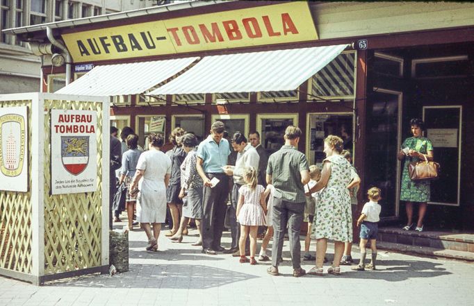 Fotografie zachycuje scénu z Východního Německa (NDR) v roce 1969. Snímek zobrazuje skupinu lidí před stánkem s nápisem "Aufbau-Tombola" na ulici Kröpelinerstrasse.