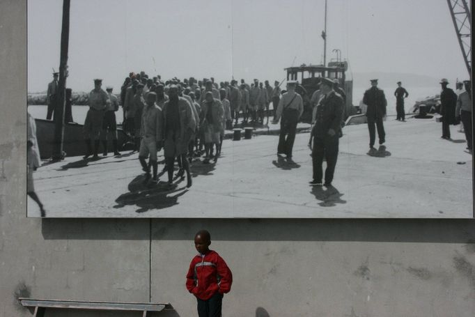 Chlapec pózuje v přístavu, kam kdysi lodě přivážely politické vězně (viz obří zvětšenina historické fotografie na zdi za ním)