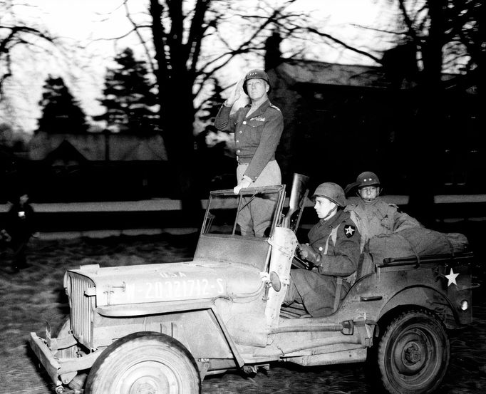 Generál Patton (stojící) zdraví vojáky Druhé pěší divize při inspekci v severní Anglii v dubnu 1944. Na zadním sedadle velitel divize generálmajor Walter M. Robertson.