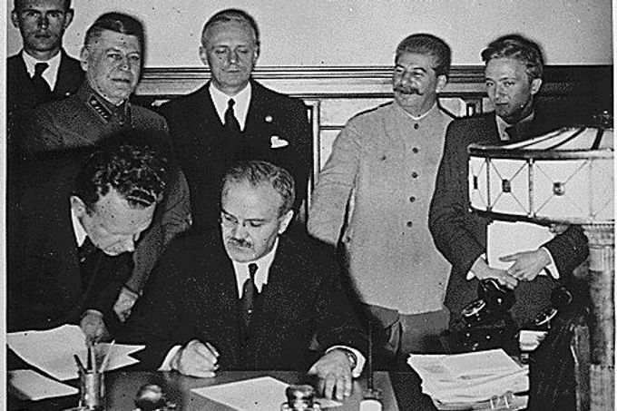 Ministr zahraničí SSSR Vjačeslav Molotov podepisuje sovětsko-německý pakt. Jeho německý protějšek Joachim von Ribbentrop s Josifem Stalinem v pozadí.