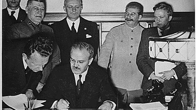 Ministr zahraničí SSSR Vjačeslav Molotov podepisuje sovětsko-německý pakt. Jeho německý protějšek Joachim von Ribbentrop s Josifem Stalinem v pozadí.