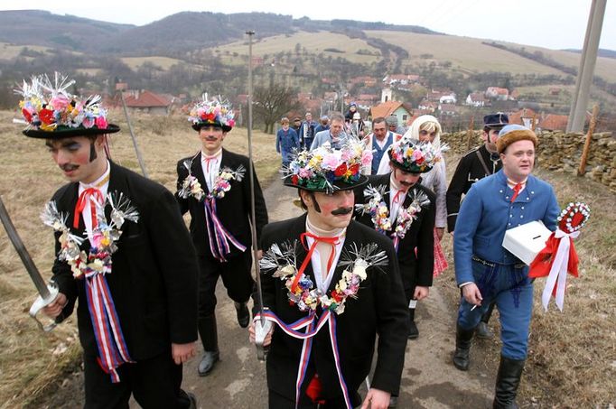 Fašankový průvod stoupá nad dědinu k bývalému JZD. Obec Komňa leží v údolí mezi kopci na okraji Bílých Karpat.