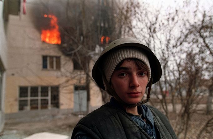 Čečenský chlapec s helmou stojí před hořící budovou během bojů o Groznyj, leden 1995. Fotografie zachycuje chaos první čečenské války.