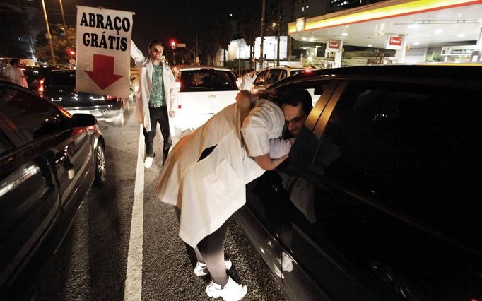 A member of the Traffic Psychologists hugs a driver next to cars along a road, while another member (L) holds a banner during their performance in Sao Paulo July 23, 2012. Traffic Psychologists is a non-profit non-governmental organization which aims to humanize traffic and reduce the level of stress caused to drivers. Sao Paulo has more than 7 million vehicles, according to figures from the state transport authority Detran. The banner reads, "Free Hug". Picture taken July 23, 2012. REUTERS/Nacho Doce (BRAZIL - Tags: TRANSPORT SOCIETY) Published: Čec. 24, 2012, 7:28 dop.