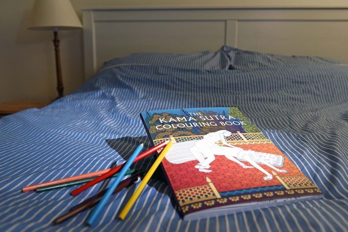 Kniha The Kama Sutra Colouring Book na posteli s pastelkami. Publikace obsahuje ilustrované erotické motivy k vybarvování, vydaná nakladatelstvím Unicorn.