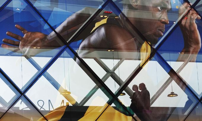 Shoppers are silhouetted through an advertisement for one of the Olympic sponsors at Westfield shopping centre near the Olympic Park at Stratford in London July 3, 2012. Sprinter Dwain Chambers, who was given a two-year doping ban in 2003, was included on Tuesday in Britain's athletics team for the London Olympics. REUTERS/Luke MacGregor (BRITAIN - Tags: SPORT OLYMPICS ATHLETICS BUSINESS) Published: Čec. 3, 2012, 4:48 odp.