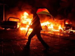 Příznivec Fatahu s palestinskou vlajkou před zapálenými auty u budovy parlamentu v Gaze
