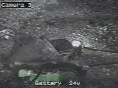 Záběr z kamery spuštěné do dolu průzkumným vrtem, zachycující část výstroje jednoho z horníků včetně důlní lampy