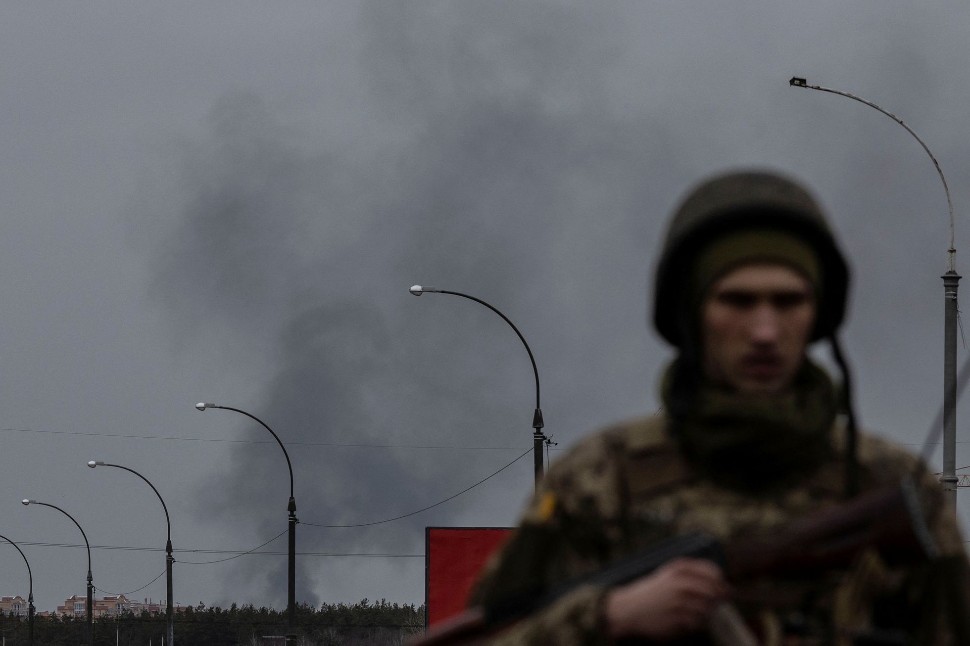 Ukrajinský voják na kontrolním stanovišti nedaleko Kyjeva. Za ním stoupá kouř z míst, ostřelovaných Rusy.