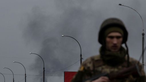 Ukrajinský voják na kontrolním stanovišti nedaleko Kyjeva. Za ním stoupá kouř z míst, ostřelovaných Rusy.