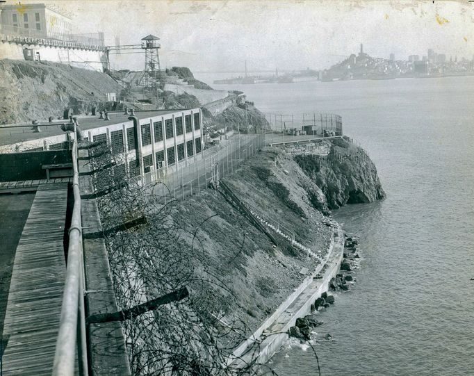 Částečný pohled na vězeňský komplex Alcatraz s ostnatým drátem, strážní věží, blokem cel a budovou dílen; v pozadí panorama San Francisca. Cca rok 1940.