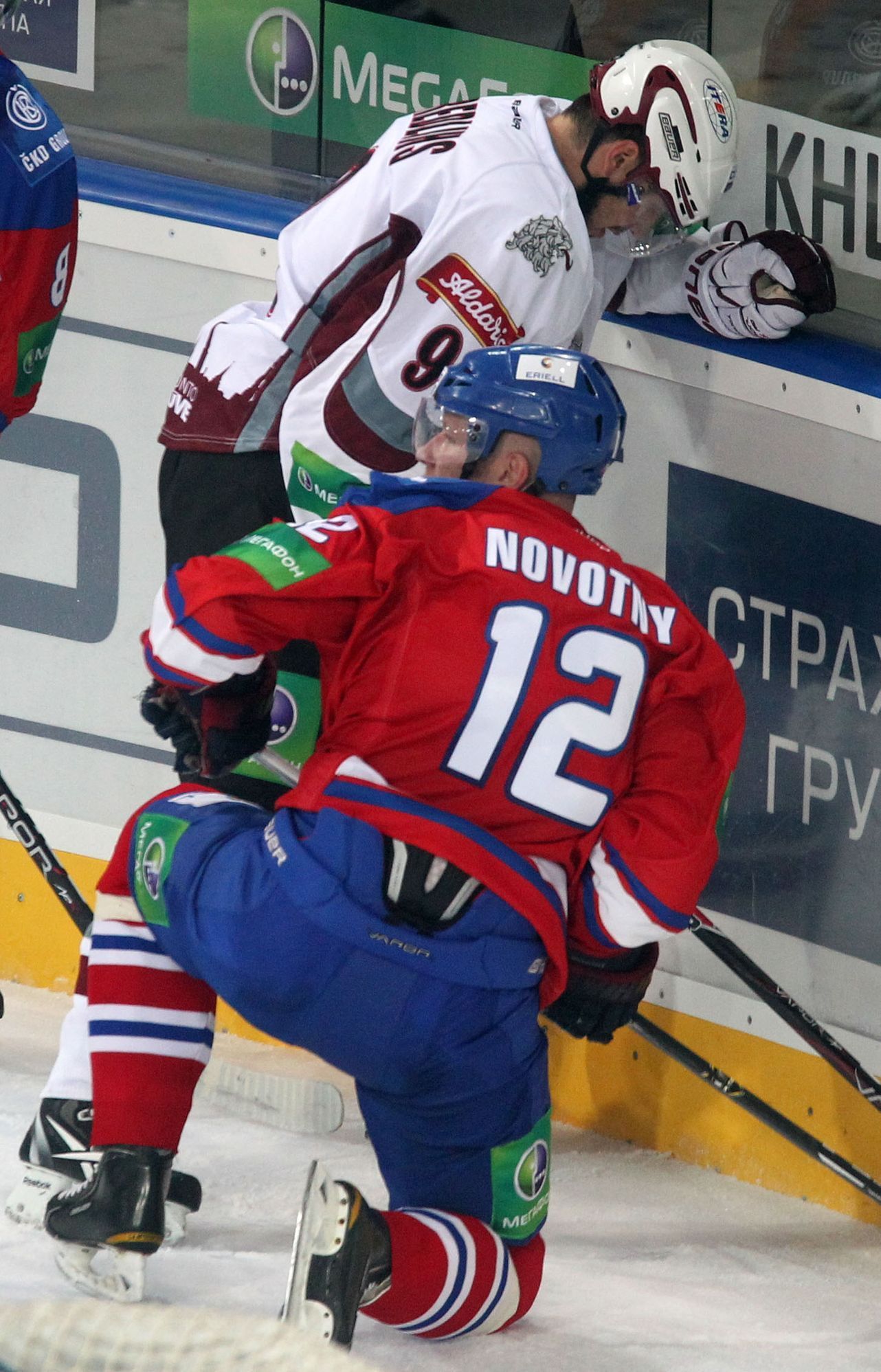 Hokejista Krisjanis Redlihs (vzadu) brání puk před Jiřím Novotným v utkání KHL 2012/13 mezi Lvem Praha s Dinamem Riga .