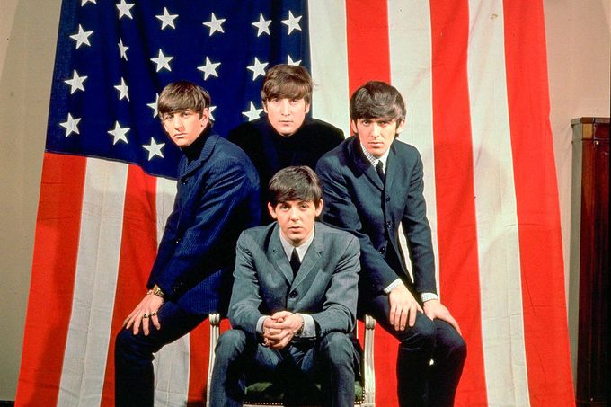 V černém saku John Lennon, pod ním Paul McCartney, vlevo Ringo Starr a vpravo George Harrison. Beatles pózují s americkou vlajkou v roce 1964