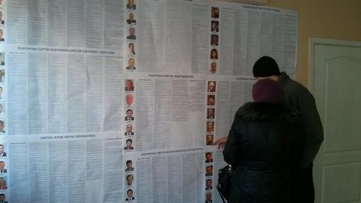 Voliči v kyjevské panelákové čtvrti Oboloň sledují kandidátní listinu ve volební místnosti.